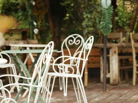 Fauteuils de fer forgé blanc aux dossiers en cœur et aux accoudoirs à volutes, autour de tables rondes, sur une terrasse de bois verdoyante