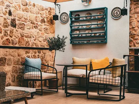 Coin de cour en pierre : deux fauteuils et une table à structure d'acier noir grillagé, coussins bleu et jaune, olivier et étagère murale