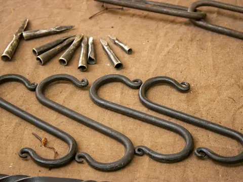 Quatre grands crochets en S forgés à la main aux extrémités enroulées, posés à côté d'embouts de métal martelé sur une toile écrue