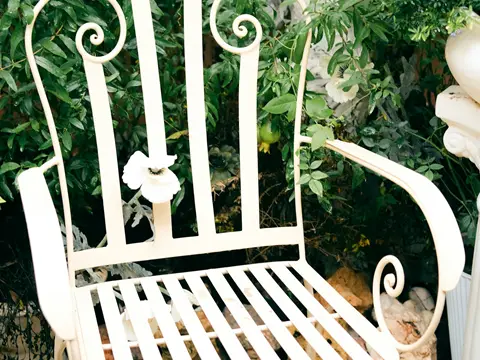 Fauteuil de fer forgé blanc à dossier à barreaux et accoudoirs à volutes, posé sur des galets au milieu des feuillages d'un jardin
