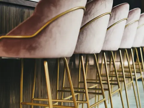 Rangée de tabourets de bar en velours rose, dossier et pieds en tube de laiton poli, devant un comptoir habillé de lattes de noyer