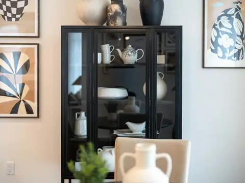 Vitrine haute à fine ossature d'acier noir et portes vitrées renfermant théière, bols et vases blancs, entre deux tableaux