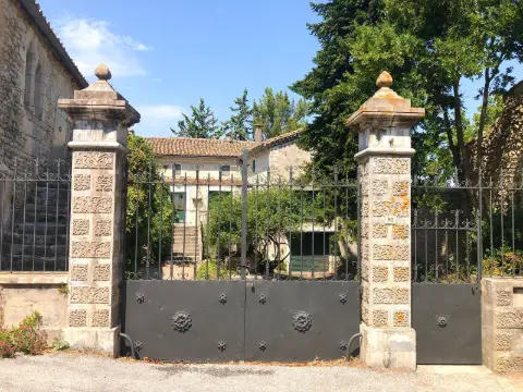 Portail battant noir à rosaces entre deux piliers de pierre sculptée, devant une bastide
