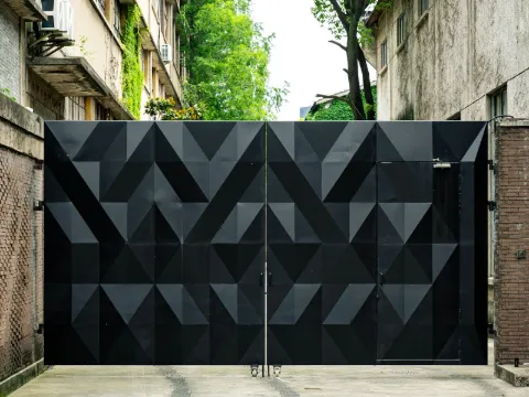Grand portail battant en acier noir mat à reliefs triangulaires, entre deux bâtiments et des arbres